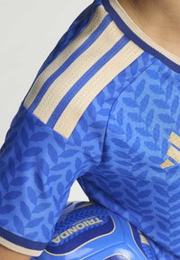 Modrá sportovní dres s listovým vzorem, béžové pruhy na ramenou, texturovaná tkanina a akcent s logem. Částečně viditelný fotbalový míč pod dres.