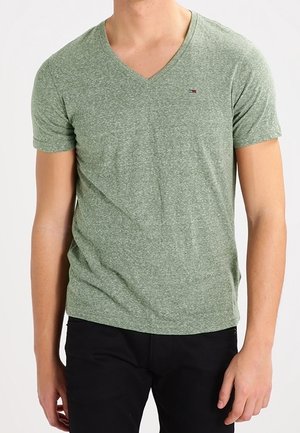 Mann trägt ein eng anliegendes grünes meliertes V-Ausschnitt-T-Shirt mit kleinem Logo auf der linken Brust und schwarze Hose, steht vor einem einfarbigen Hintergrund.