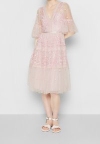 Robe en tulle rose avec un décolleté en V, des manches évasées, des détails floraux brodés et une jupe superposée, assortie à des sandales blanches.