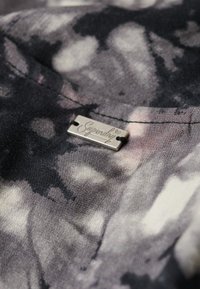 Koszulka z krótkim rękawem w czarno-szary abstrakcyjny wzór, z metaliczną srebrną metką logo przymocowaną przy dekolcie.