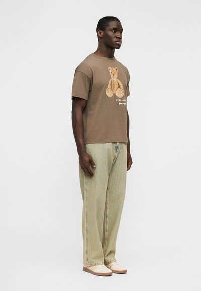 Καφέ βαμβακερό μπλουζάκι με γραφικό αρκουδάκι και κείμενο, σε συνδυασμό με ανοιχτά πράσινα παντελόνια φαρδιάς γραμμής και μπεζ υποδήματα.