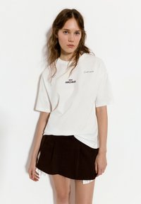 Valkoinen ylisuuri t-paita, jossa on mustalla painettu "NEW ARCHIVE". Yhdistettynä lyhyeen ruskeaan laskostettuun hameeseen, jossa on frangeilla viimeistellyt reunat.