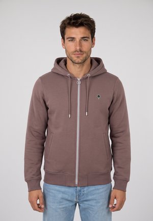 Hombre de pie con sudadera marrón con cremallera con un pequeño logo de árbol y vaqueros azul claro sobre un fondo liso.