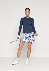 Top da golf a maniche lunghe blu navy con zip a metà, abbinato a una gonna bianca con un motivo astratto blu. Il modello tiene una mazza da golf e indossa scarpe bianche.