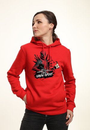 Donna con una felpa con cappuccio rossa di Deadpool che guarda di lato con le mani nelle tasche, testo sulla felpa "Change Forever" con design grafico.