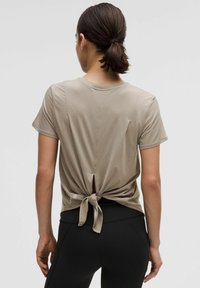 Beige short-sleeve T-shirt with a tied back detail and cutout. Soft fabric, relaxed fit, and a subtle logo at the lower back.