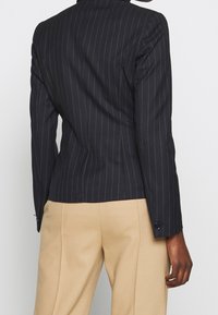 Osoba w dopasowanej czarnej marynarce w prążki i beżowych spodniach, widoczna z tyłu na jednolitym jasnym tle.