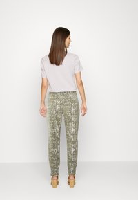 T-shirt cinza claro combinado com calças verde-oliva texturizadas com um padrão irregular brilhante. Modelo visto de costas, em pé sobre um fundo neutro.