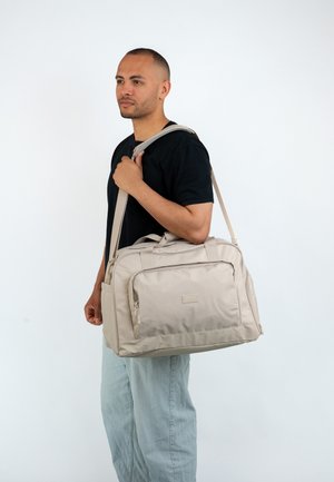 Beige Reisetasche mit einer glatten, strukturierten Oberfläche, doppelten Griffen und einem abnehmbaren Schultergurt. Verfügt über eine vordere Reißverschlusstasche und ein Logo-Detail.