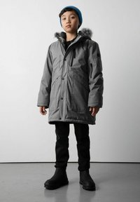 Dziecko ubrane w szary zimowy płaszcz z futrzanym kapturem, czarne spodnie, czarne buty oraz niebieski pleciony beret, stojące na szarym podłodze przy gładkiej ścianie.