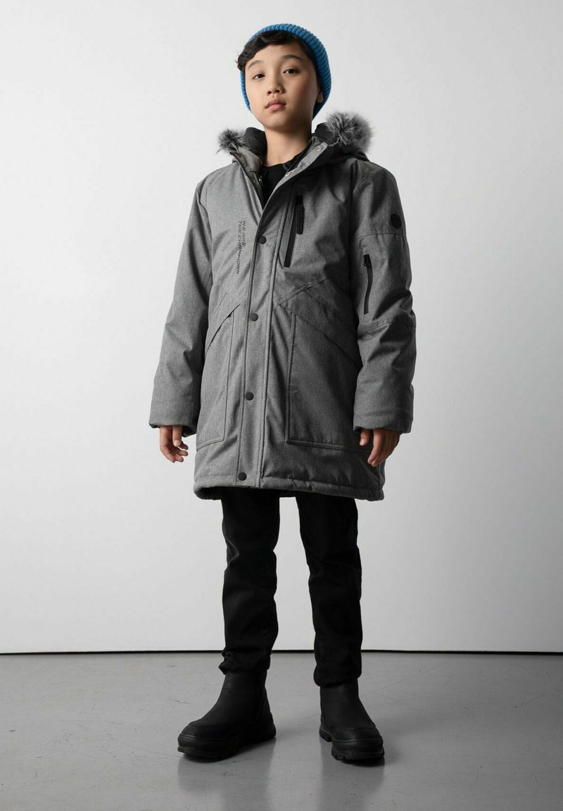 Dziecko ubrane w szary zimowy płaszcz z futrzanym kapturem, czarne spodnie, czarne buty oraz niebieski pleciony beret, stojące na szarym podłodze przy gładkiej ścianie.