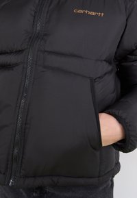 Svart pufferjakke med glidelås, med en liten Carhartt-logo i oransje og side lommer med en litt blank tekstur.