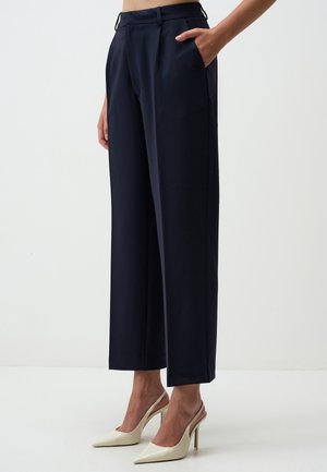 Woman wearing high-waisted, wide-leg navy trousers with side pockets and white pointed-toe high heel slingback shoes.