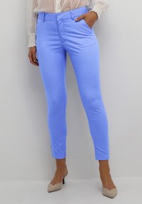 Vrouw in een strakke lichtblauwe broek en beige hoge hakken, staand tegen een effen lichte achtergrond.