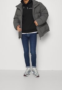 Giacca piumino grigia con cappuccio sopra una felpa nera, abbinata a jeans skinny blu e sneakers grigie con dettagli rossi e blu.