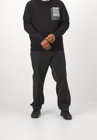 Schwarzer Pullover mit einer quadratischen Tasche, die ein graues Calvin Klein-Logo zeigt, kombiniert mit dunkelgrauer Hose und schwarzen Schuhen, schlichtes Design.