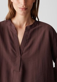 Woman wearing a loose, long-sleeve, dark brown blouse with a split neckline and collarless design, shown from the shoulders to mid-chest.