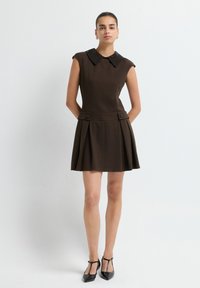 Frau in einem ärmellosen dunkelbraunen Kleid mit schwarzem spitzem Kragen, plissiertem Rock und schwarzen T-Strap-Absätzen, die mit den Händen hinter dem Rücken steht.