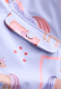 Gros plan sur un tissu violet pastel avec impression licorne et fermeture par bouton-pression sur un petit rabat de poche.