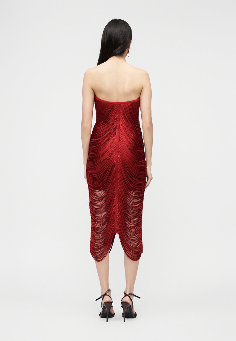 Femme aux longs cheveux bruns portant une robe midi rouge froncée sans bretelles et des sandales à talons hauts noires, debout de dos sur un fond uni.