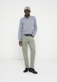 Γκρι φούτερ με φερμουάρ, ανοιχτοπράσινο παντελόνι και μαύρα παπούτσια. Το καπέλο είναι μπεζ με λογότυπο. Το σύνολο έχει casual και εφαρμοστή σχεδίαση.