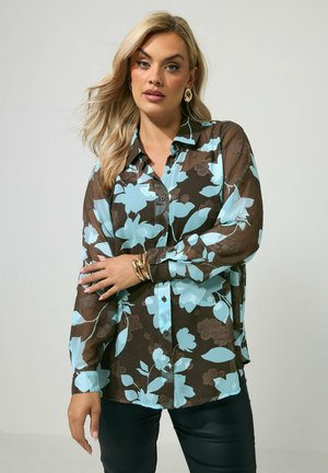 Vrouw draagt een doorzichtige bruine blouse met knopen en grote blauwe bloemprint, zwarte broek, gouden oorbellen in de vorm van hoepels en gouden armbanden, tegen een effen achtergrond.