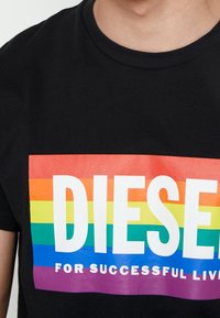 Mustat puuvillapaitat, jossa on värikkäitä sateenkaariraidoilla varustettu suorakulmainen graafinen kuvasto. "DIESEL" ja "MENESTYVÄN ELÄMÄN PUOLESTA" painettuna paksuilla valkoisilla kirjaimilla.