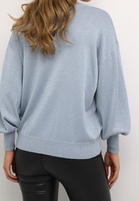 Femme aux longs cheveux ondulés portant un pull bleu clair scintillant avec des manches bouffantes et un pantalon en cuir noir ajusté, vue de dos.