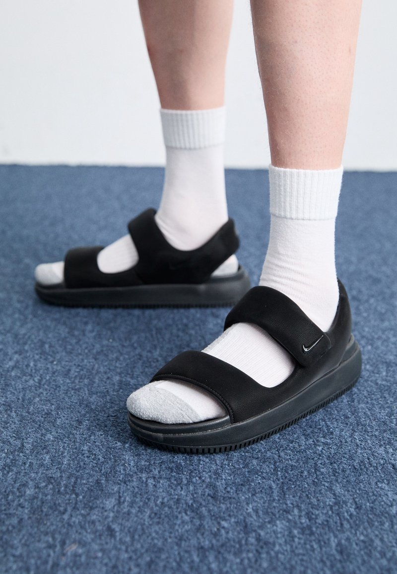 Piernas con calcetines blancos hasta la altura del tobillo y sandalias negras de Nike, de pie sobre un suelo alfombrado azul.