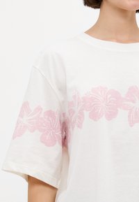 Witte katoenen t-shirt met een roze hibiscus bloem print langs de zoom. Korte mouwen met een relaxte pasvorm; zachte textuur.