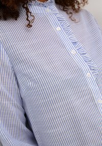 Chemise blanche à rayures verticales bleues, avec un devant à boutons, un col volanté et un tissu lisse et léger.