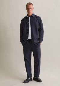 Marineblauw overhemd met knopen en een borstzak, gedragen over een wit T-shirt. Gecombineerd met bijpassende marineblauwe broek en zwarte lederen instappers.