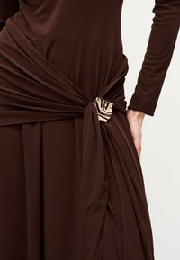 Καφέ φόρεμα με μακριά μανίκια, μαζεμένο και σφιχτά δεμένο στη μέση με μια υφασμάτινη χρυσή ορθογώνια αγκράφα.