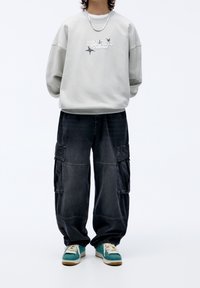 Persona che indossa un maglione grigio chiaro con testo, pantaloni cargo neri, sneakers turchesi e una collana in argento, in piedi contro uno sfondo bianco.