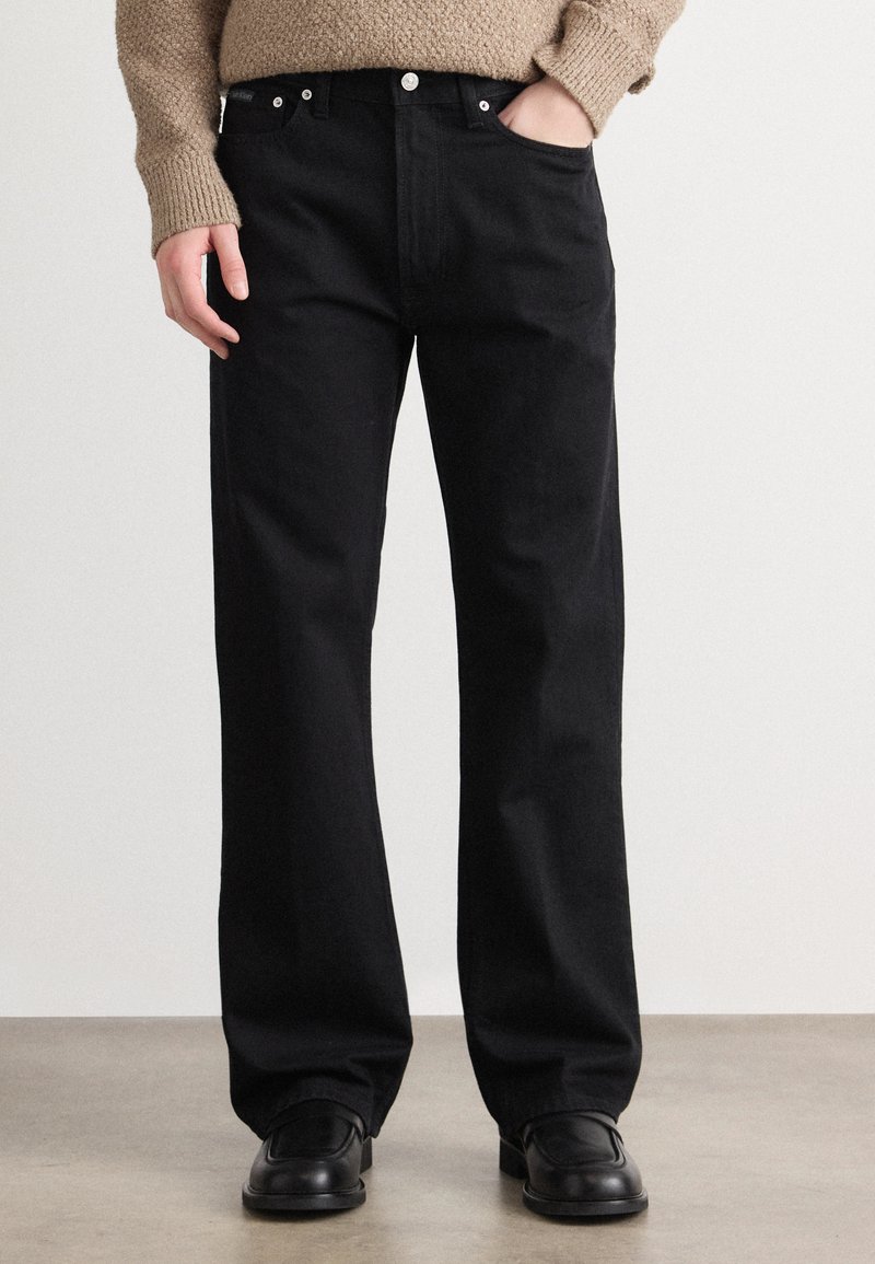 Juodi džinsai su tiesiomis kojomis, penkios kišenės, metaliniai elementai ir subtilus tekstūravimas. Suderinti su juodais batais.