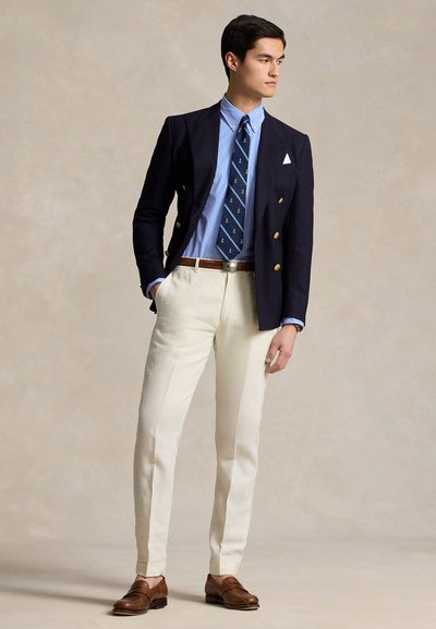 Navy double-breasted blazer, light blue striped shirt, patterned tie, cream tapered trousers, and brown leather loafers. Plain background.