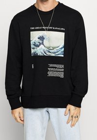 Sudadera negra con un gráfico frontal de "La Gran Ola de Kanagawa" y texto debajo. Tela suave, ajuste relajado, puños y dobladillo acanalados.