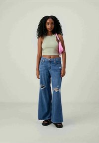 Jauna moteris su garbanotais plaukais, vilkinti žalią trumpą viršutinę dalį, suplyšusius plačius džinsus, juodas šlepetes ir nešanti rožinę petinę rankinę.