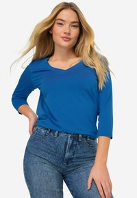 Modrá, dlouhá halenka s výstřihem do V, vyrobená z měkké tkaniny, v kombinaci s pruhovanými džíny s vysokým pasem. Halenka má volný střih a hladkou texturu.