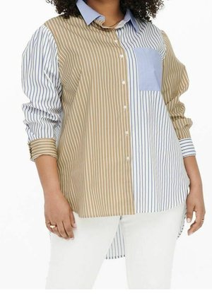 Vrouw draagt een lange mouwen, knoopjes overhemd met gemengde verticale strepen in beige, blauw en wit, gecombineerd met een witte broek.