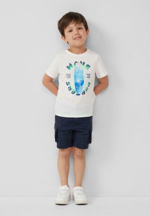 Biała koszulka z krótkim rękawem z kolorową grafiką deski surfingowej i napisem, zestawiona z granatowymi szortami cargo i białymi sneakersami.