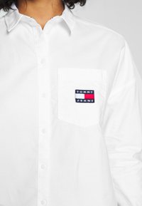 Vit skjorta med knapp uppe, tillverkad av bomull, med en vänster bröstficka som har ett Tommy Jeans logotypmärke i marinblått, rött och vitt.