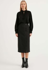 Svart långärmad blus med knytkrage, matchad med en svart pencilkjol med randigt mönster som har sidofickor och knappar framtill. Svarta klackstövlar.