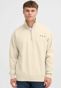 Beige hoodie met halve rits in een relaxed fit, met ribgebreide manchetten en onderkant, en zwarte "A.R.C." letters op de linkerborst. Zachte textuur.