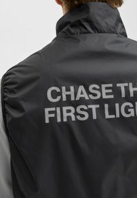 Zwarte geïsoleerde vest met een hoge kraag, grijze accenten en de tekst "CHASE THE FIRST LIGHT" in vette, reflecterende letters over de achterkant.
