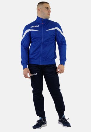Mand iført en blå og hvid sportstrøje med lynlås og sorte sportbukser, der står med hovedet vendt mod højre på en hvid baggrund.