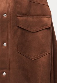 Καφέ σατέν μπουφάν με εμπρόσθια τσέπη με καπάκι και ασημένια κουμπιά πιέτας σε μονόχρωμο φόντο.