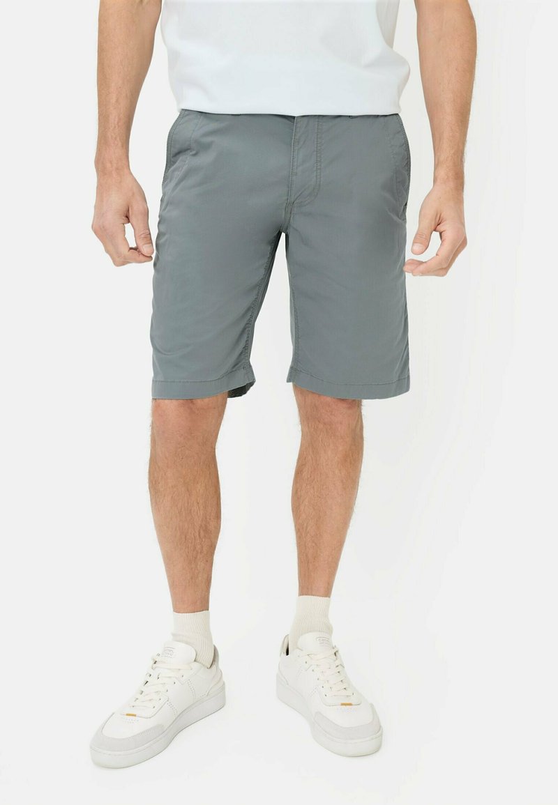 Mann trägt graue knielange Shorts, weiße Sportsocken, weiße Turnschuhe und ein weißes T-Shirt, steht mit locker an den Seiten hängenden Armen.