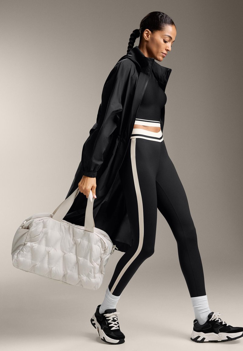 Fekete hosszú kabát, testhezálló fekete felső, magas derekú fekete leggings krémszínű csíkokkal, valamint texturált fehér duffel táska fogantyúkkal. Fekete sportcipők.