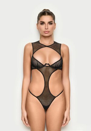 Mujer vestida con un body de malla negro con cortes, cuello alto y parte trasera tipo tanga, de pie contra un fondo claro y liso.